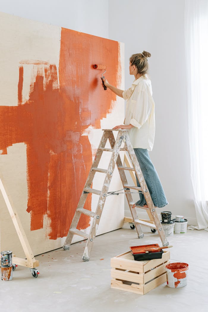 A woman stands on a stepladder, painting a rooms wall during home renovation.